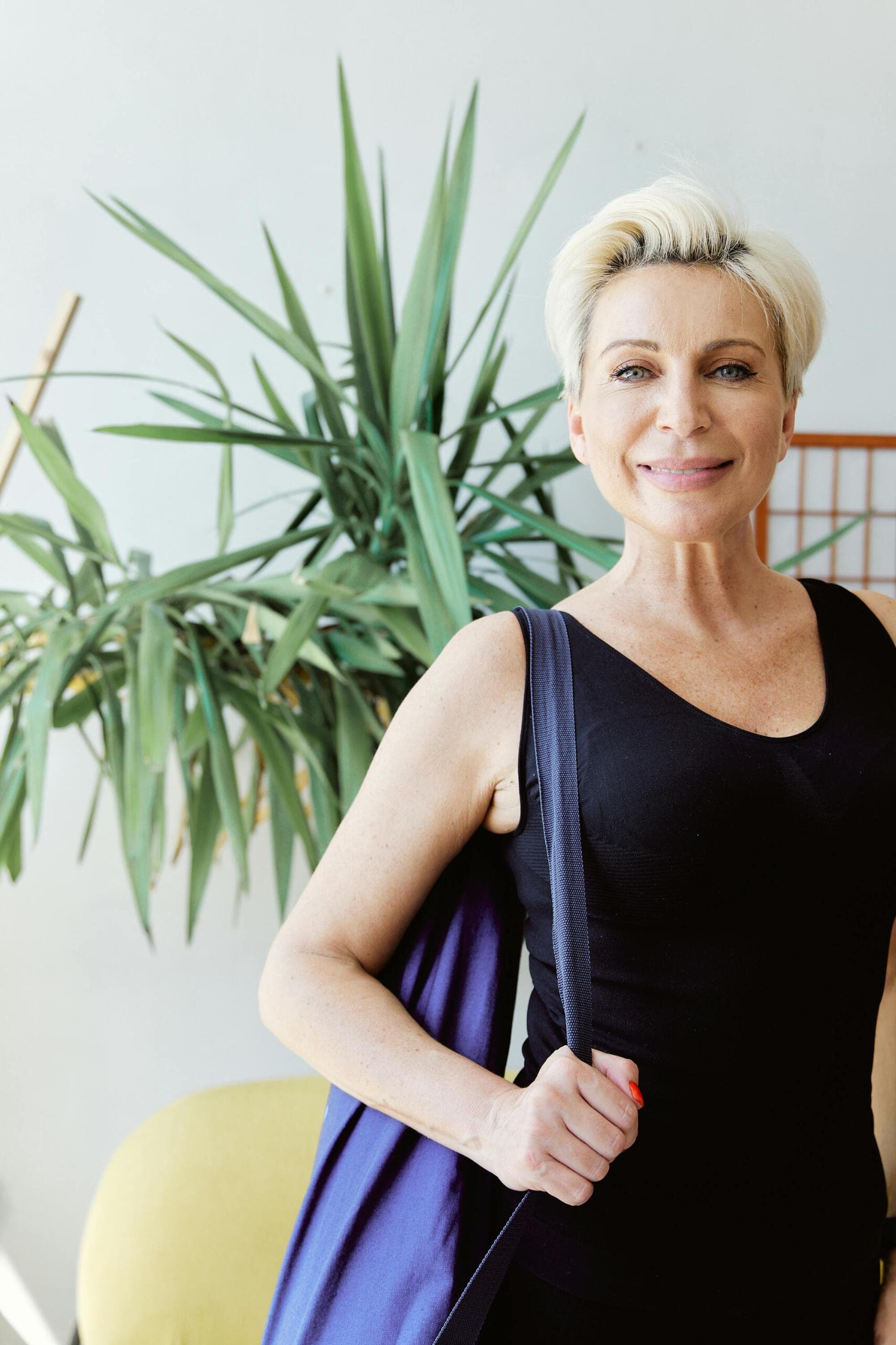 A smiling older woman with short blonde hair carrying a yoga bag, standing in front of a large houseplant.